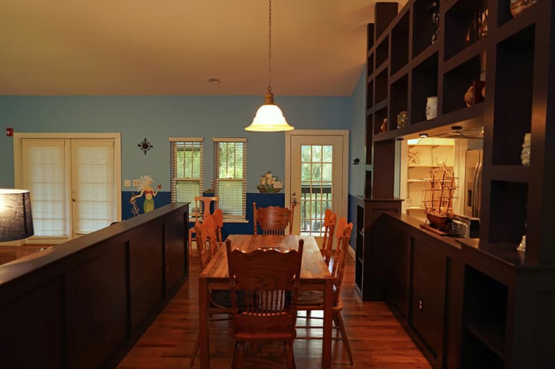 This inviting dining area features a long wooden table with matching chairs, set between a dark wooden built-in unit and a counter, with light blue walls and hardwood floors.