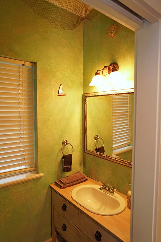 This inviting bathroom features vibrant green textured walls, a wooden vanity with a white sink, a mirror, and a window with blinds, creating a warm ambiance.