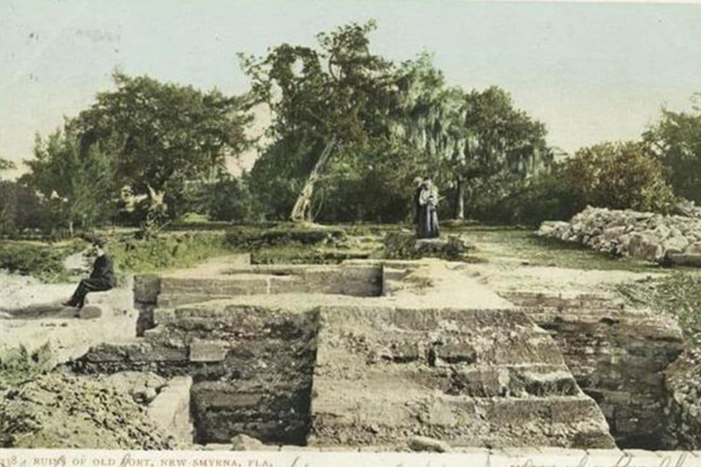 A historic, colored photograph of the ruins of Old Fort in New Smyrna, Florida, featuring two figures in the background and a seated individual in the foreground.