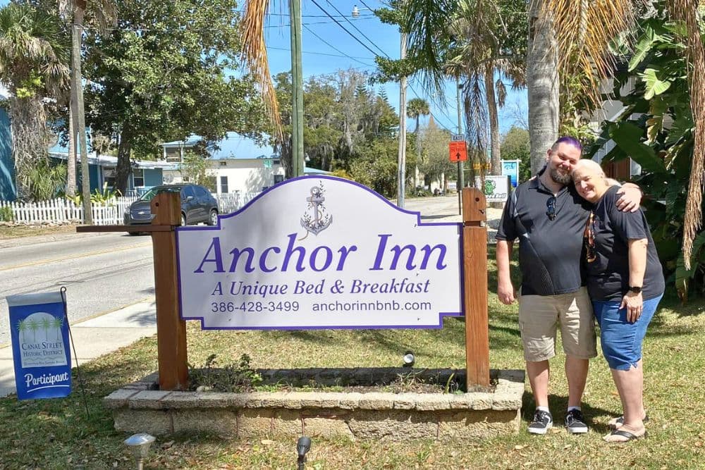 A smiling couple poses beside the Anchor Inn Bed & Breakfast sign in a sunny outdoor setting.