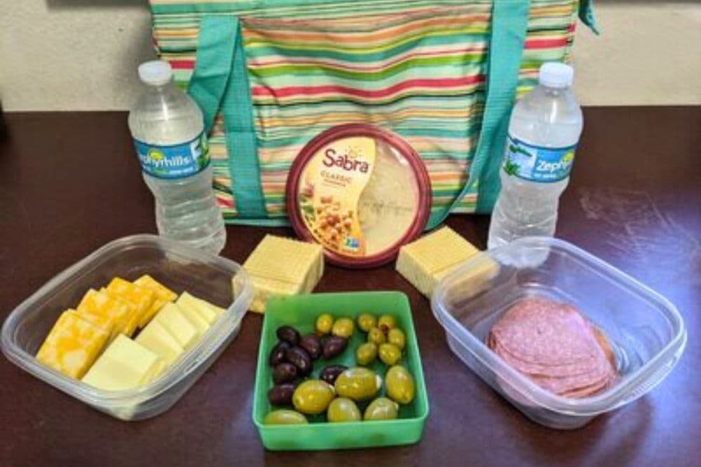 A picnic spread featuring a striped bag, water bottles, cheese, salami, olives, crackers, and hummus.