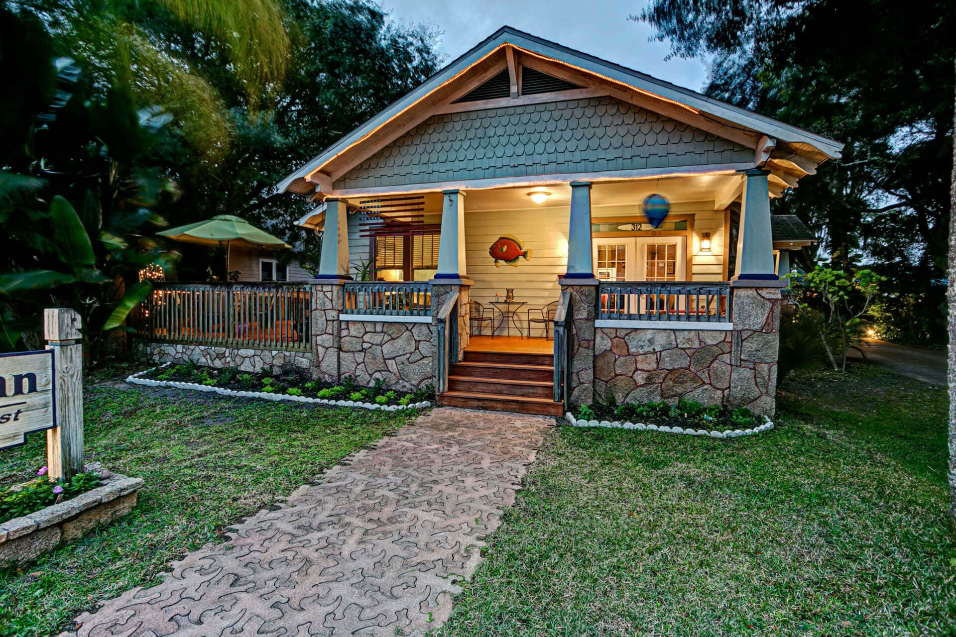 Daytime exterior of the Anchor Inn featuring stone siding on the porch and green shingles with a walkway leading to the porch to sit on a patio set with 2 chairs and a table with a umbrella.