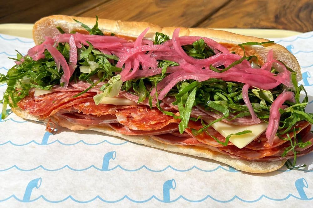 A sub sandwich filled with layers of salami, cheese, greens, and pickled onions on a soft hoagie roll.