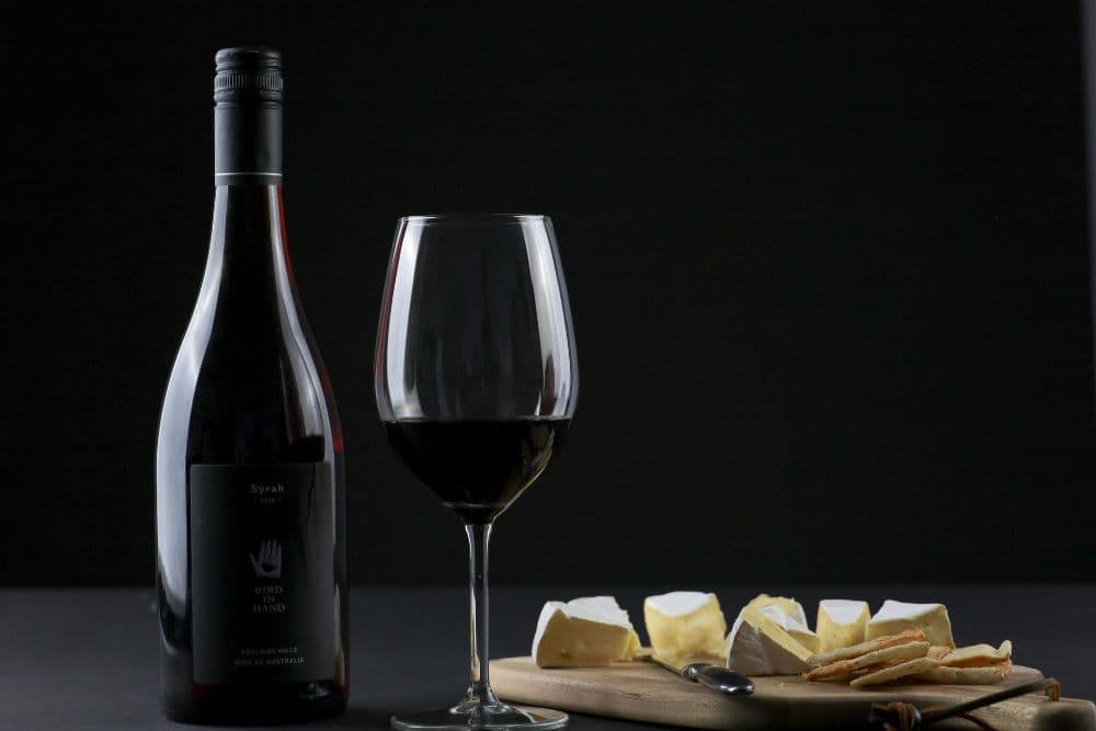 A bottle of Syrah wine, a glass of red wine, and a platter of cheese with crackers are set against a dark background.