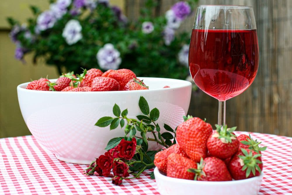 A bowl of fresh strawberries sits beside a glass of red drink on a checkered tablecloth, with flowers in the background.