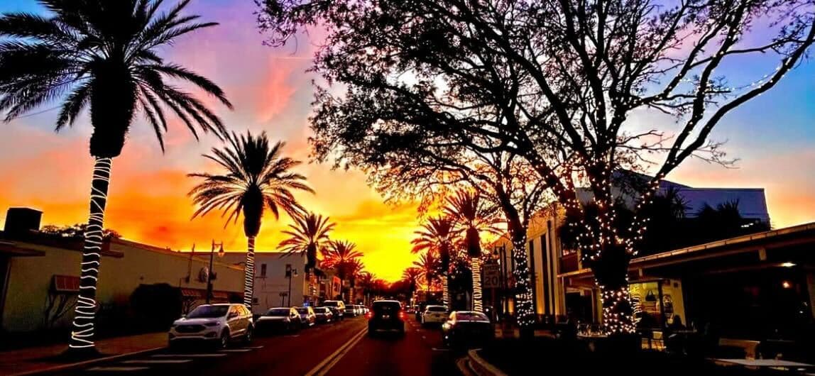 A vibrant sunset illuminates a palm-lined street adorned with holiday lights.