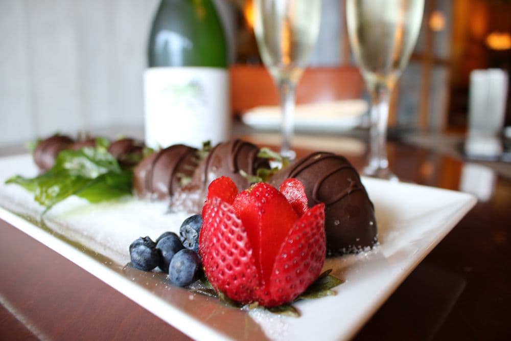 A plated dessert featuring chocolate-covered strawberries, a carved strawberry flower, blueberries, and a bottle of champagne in the background.