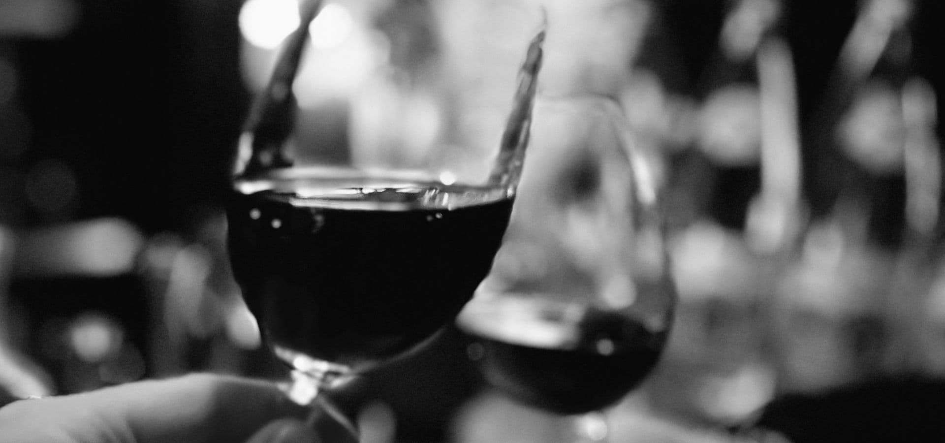 A close-up of a hand holding a wine glass, with blurred glasses in the background, all captured in black and white.