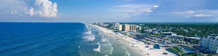 A panoramic view of a beach with waves, shoreline, and coastal buildings under a clear blue sky.