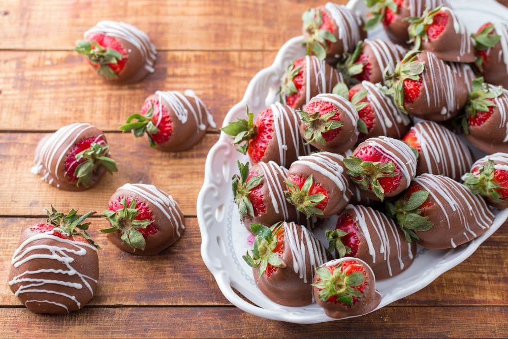 A platter of chocolate-covered strawberries drizzled with white chocolate, set on a wooden table.