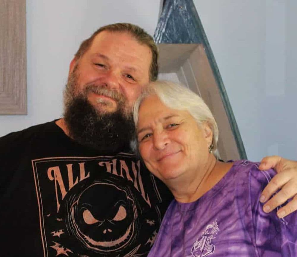 A smiling man with a beard and a woman with short gray hair embrace in a cozy setting.