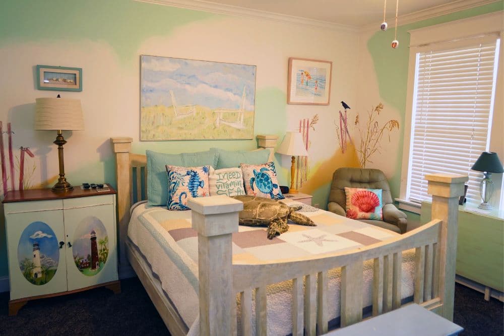 A cozy bedroom featuring a bed with decorative pillows, a sea-themed quilt, art on the walls, and colorful accents.