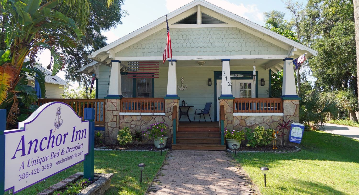 Front view of a quaint home with porch, white columns, flowers, palm trees and sign in the front yard