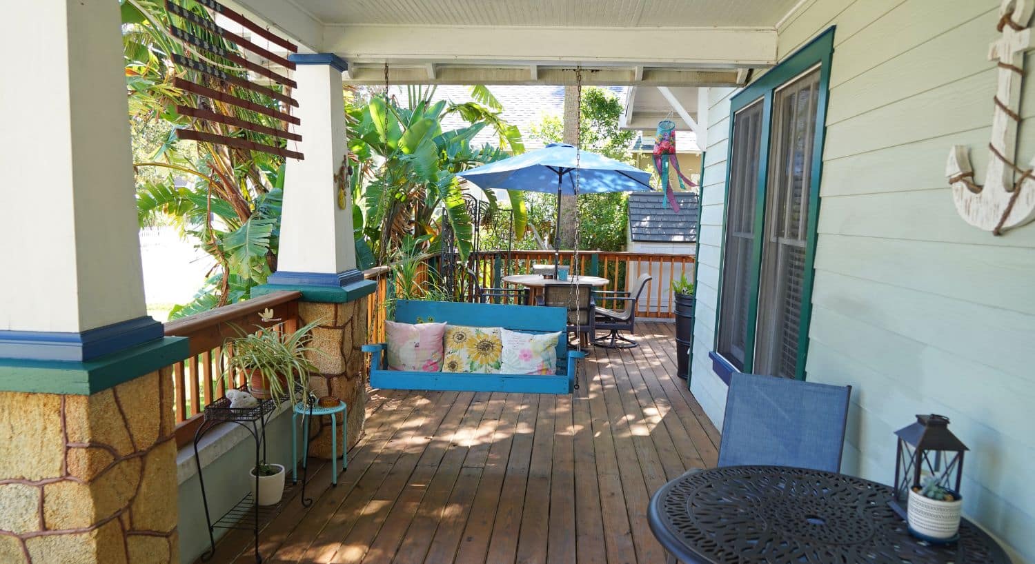 Front porch of a home with swing chair, table with chair, patio table with umbrella and lush plants and palm trees