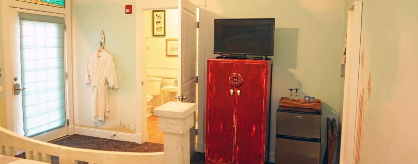 Bedroom with white bed, tall dresser with TV, French door with stained glass window above and doorway into attached bath