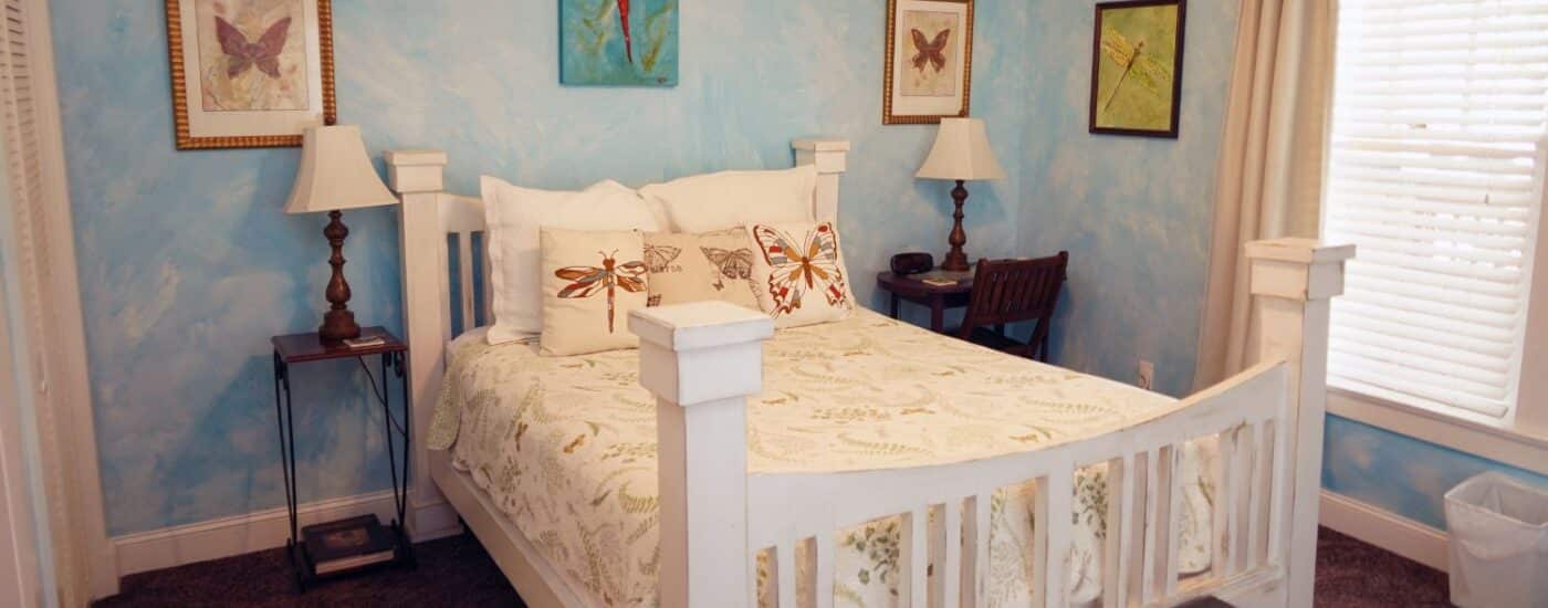 Bedroom with white queen bed, side tables with lamps, artwork, window with blinds and white curtains