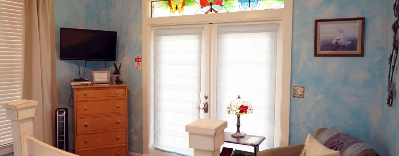 Bedroom with French doors, decorative butterfly stained glass window, plush sitting chair, dresser with TV