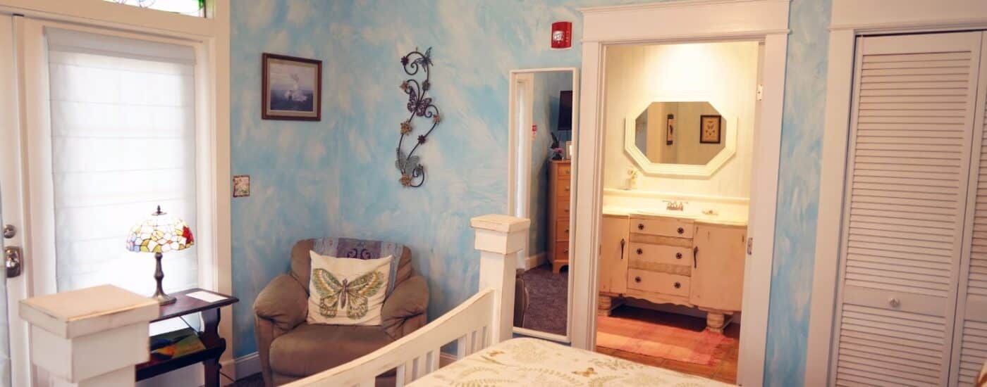 Bedroom with white queen bed, plush sitting chair, French door with blinds and doorway into attached bath