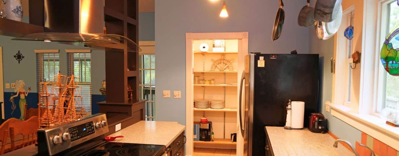 Galley kitchen area with black and stainless steel appliances, blue walls, hanging pot rack and doorway to a pantry wth shelving