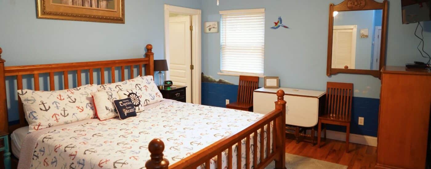 Bedroom with king bed, quilt in anchor pattern, dropleaf table with two chairs, dresser with TV