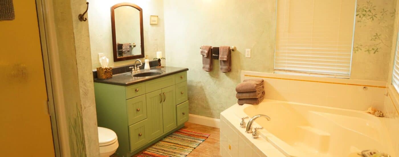 Large bathroom with green vanity, mirror, toilet, plush brown towels, corner soaker tub