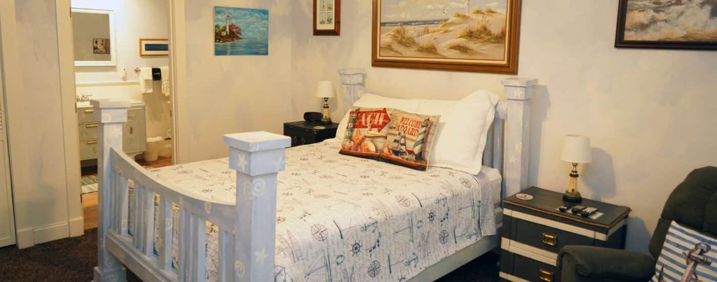 Bedroom with queen bed, seashore artwork, plush sitting chair and doorway into attached bathroom