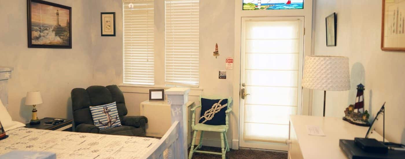 Cozy bedroom with queen bed, plush sitting chair, dresser, lamp and door with stained glass window above