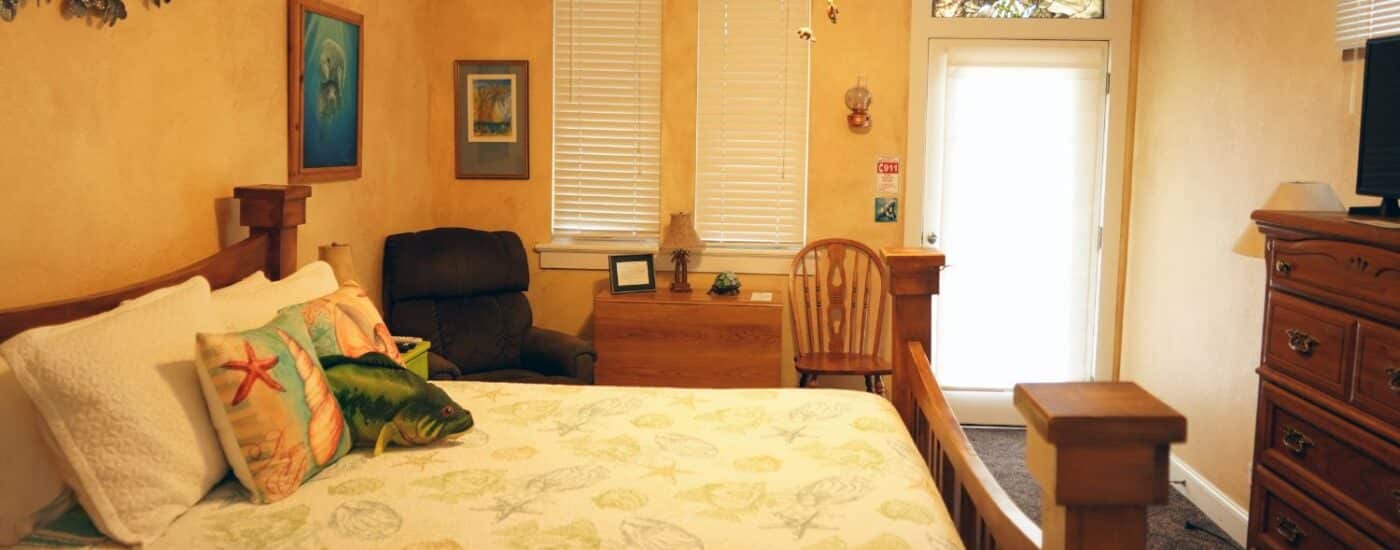 Bedroom with seashell pattern quilt on queen bed, sitting chair, dresser with TV and door with stained glass window above