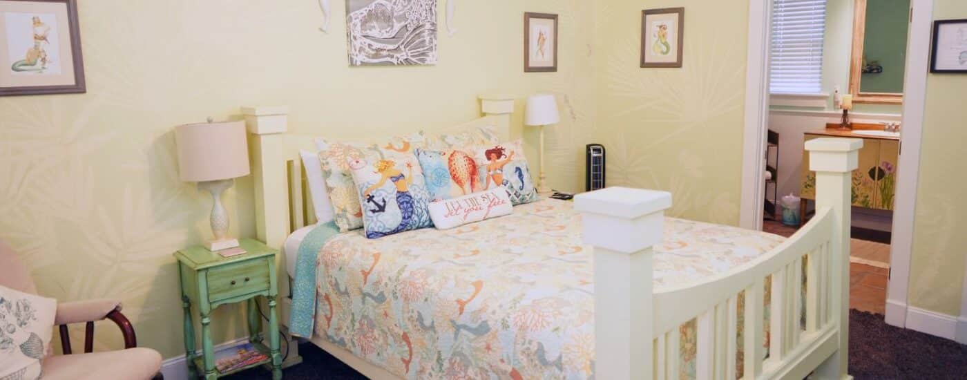 Bedroom with white queen bed, rocking chair, side tables with lamps, colorful artwork and doorway into attached bath