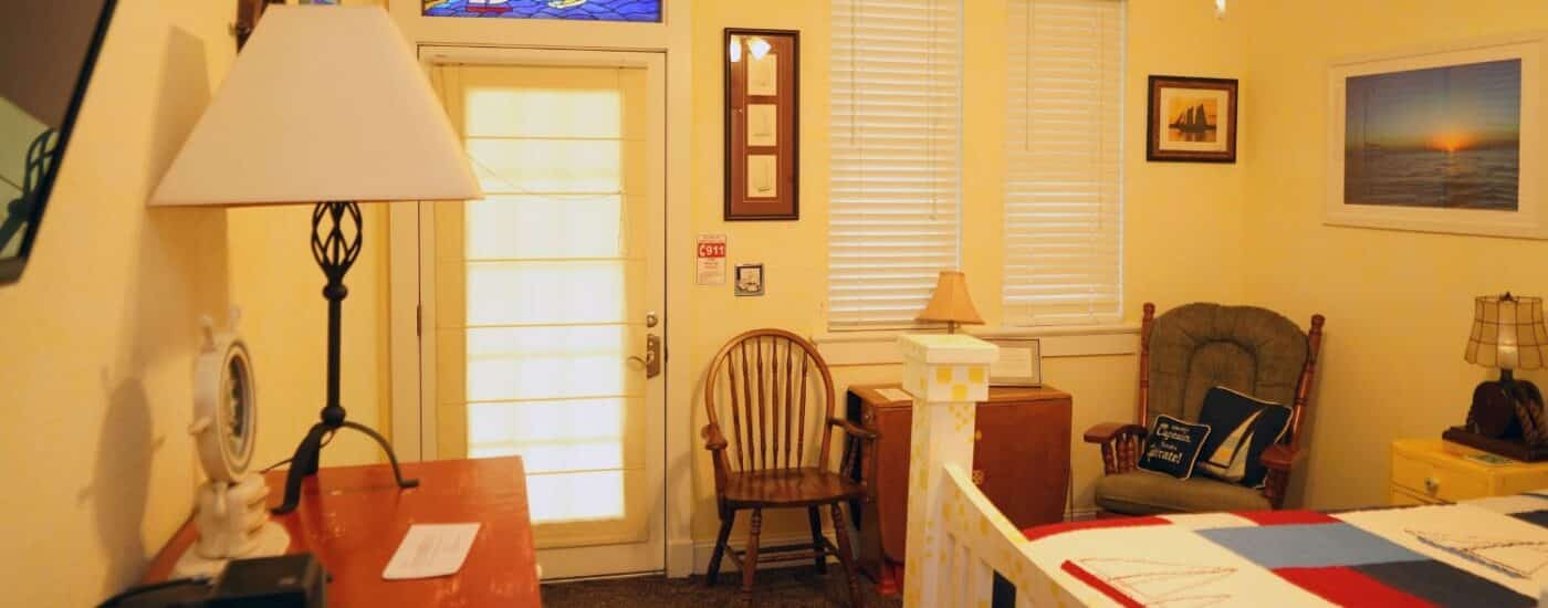 Bedroom with queen bed in patchwork quilt, rocking chair, dresser with lamp and door with stained glass window above