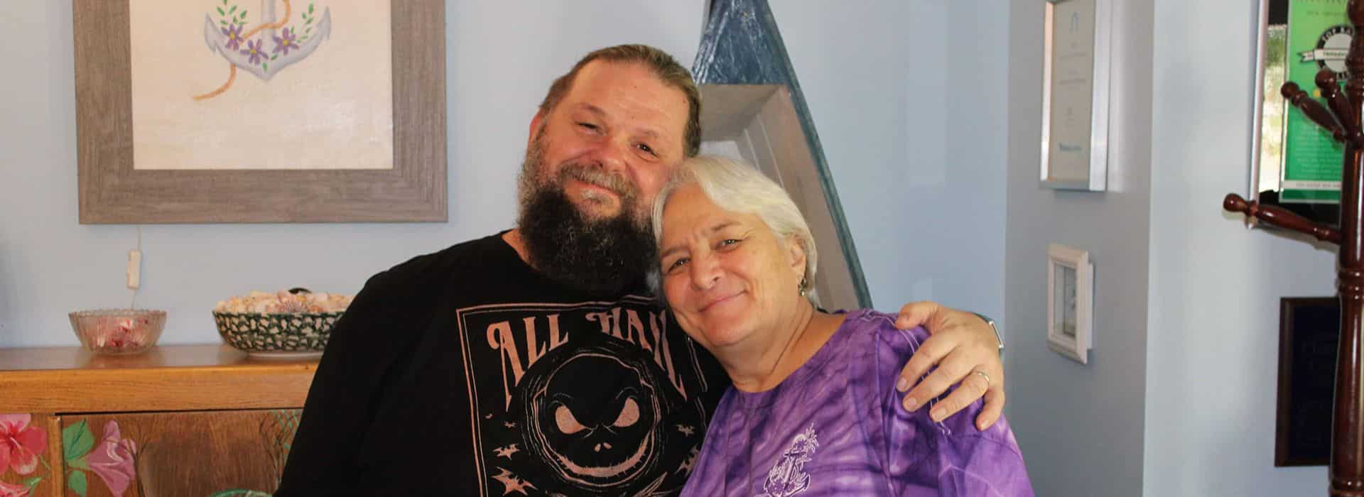 A man in a beard and black shirt hugging a woman in a purple shirt in a home