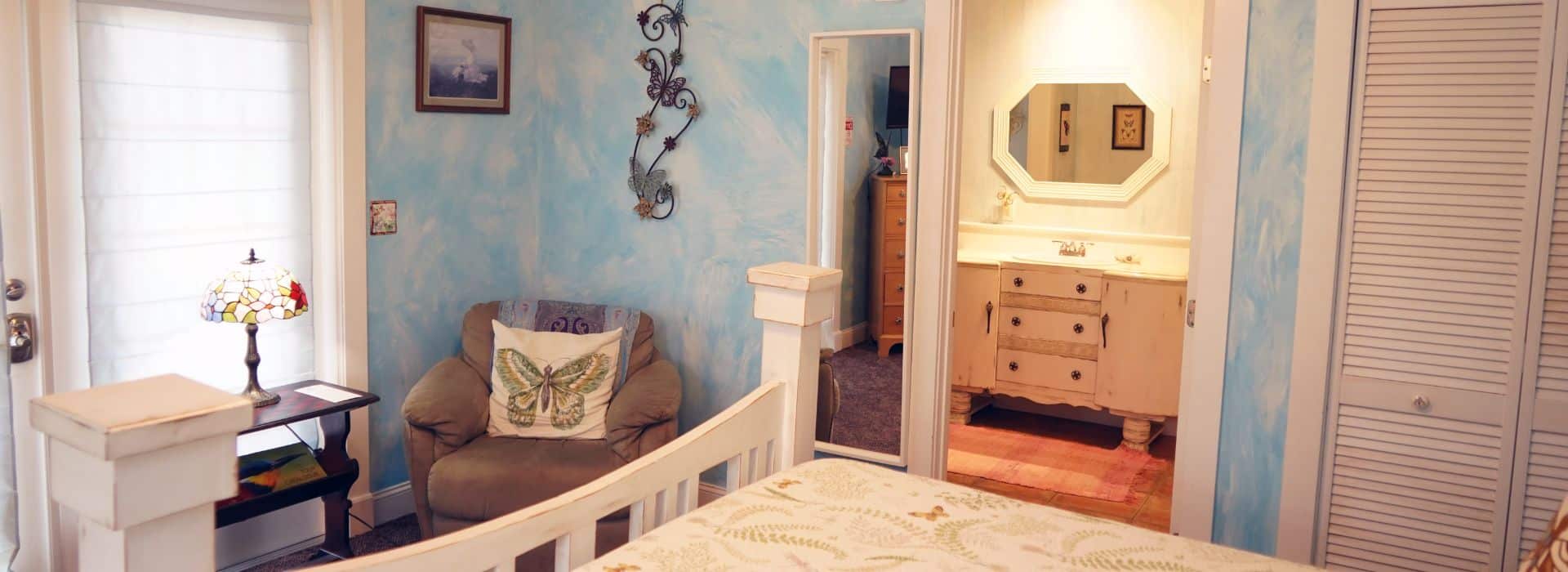 Bedroom with white queen bed, plush sitting chair, French door with blinds and doorway into attached bath