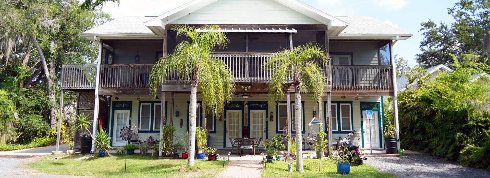 Back view of a home with large patio, upper deck, lush bushes and two tall palm trees