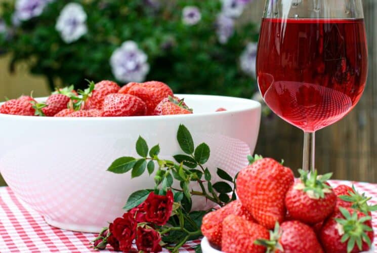 A large white bowl of fresh strawberries next to a glass of red wine on a table with a red and white checkered cloth