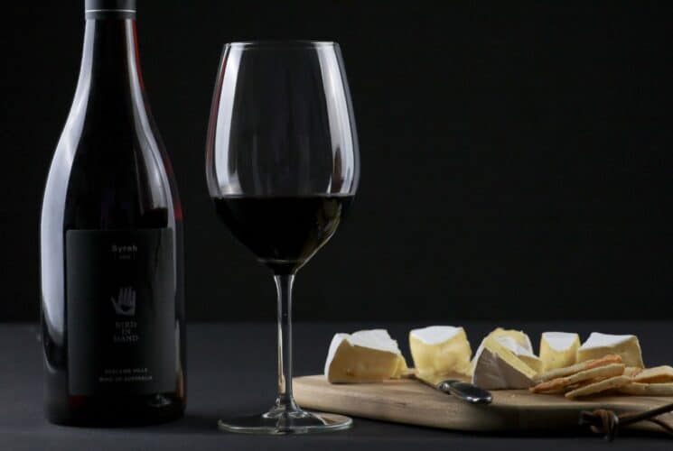 A bottle and glass of red wine next to a wood board with sliced cheese and crackers