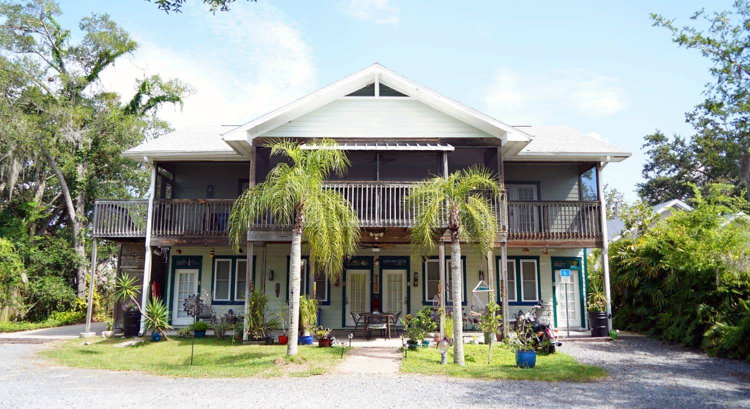 Back view of a two story home with upper level decking, lush green plants, palm trees and gravel drive