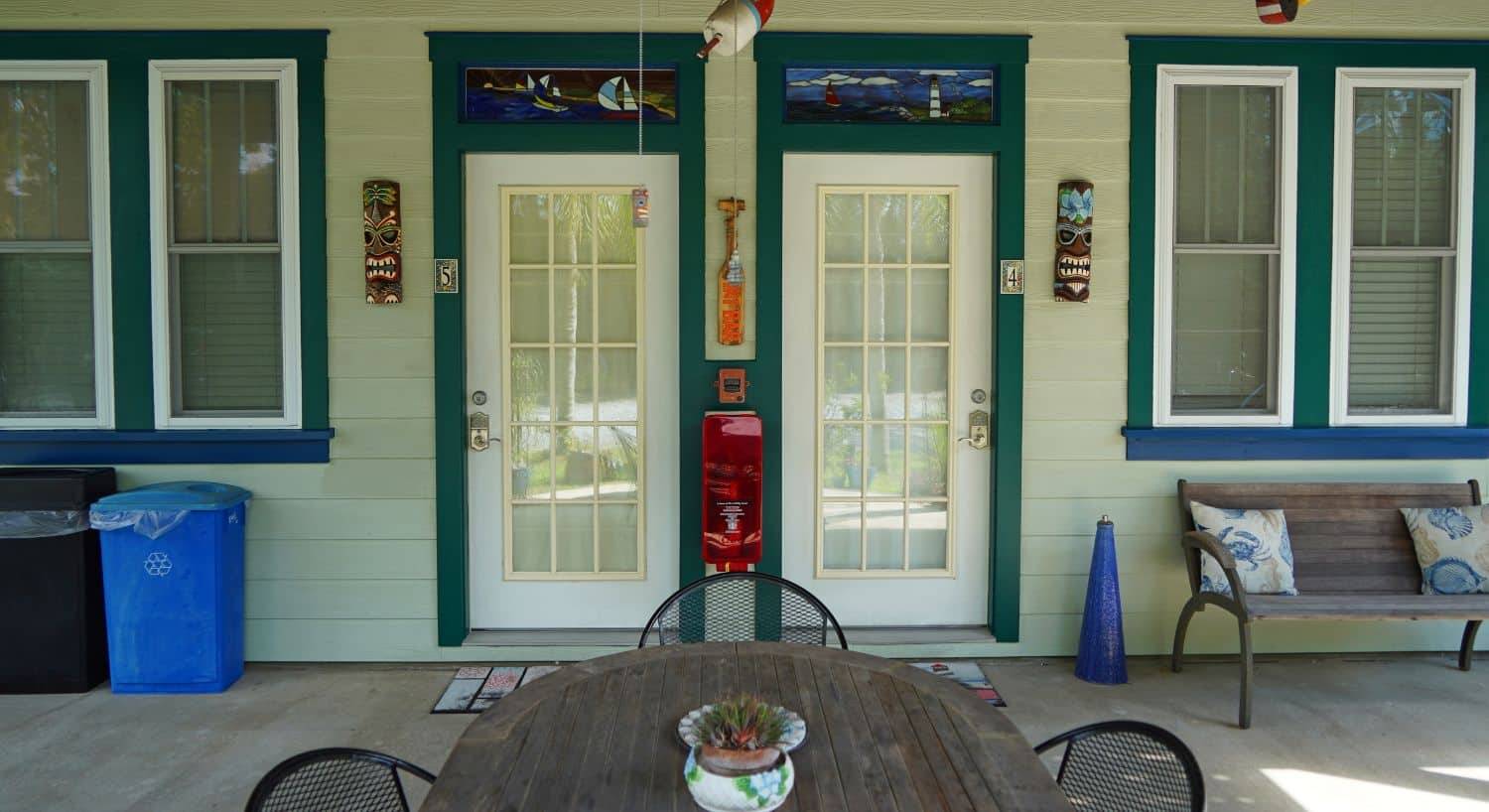 Outside porch of a home with two doors, for windows, wood table with black chairs and wood bench